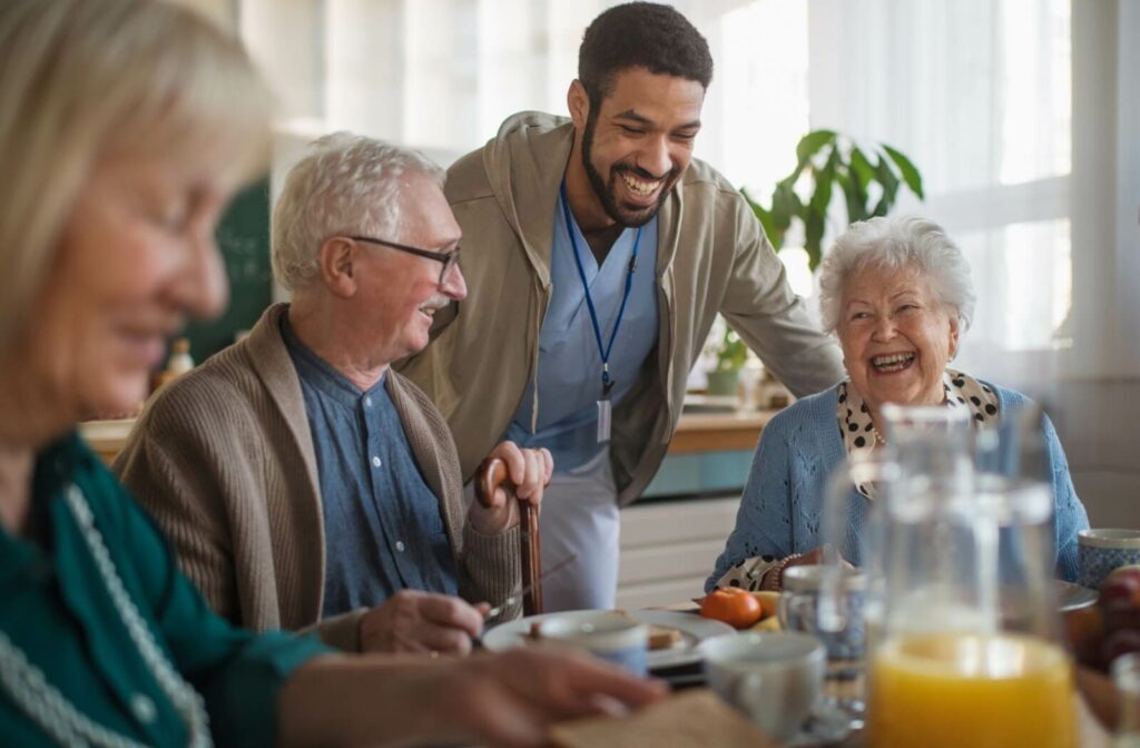 A smiling caregiver checks in on 3 residents during breakfast in memory care