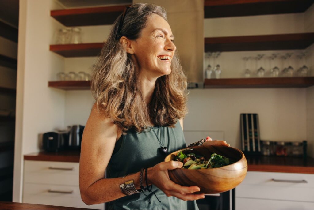 A smiling older adult woman holding a wooden bowl full of salad.